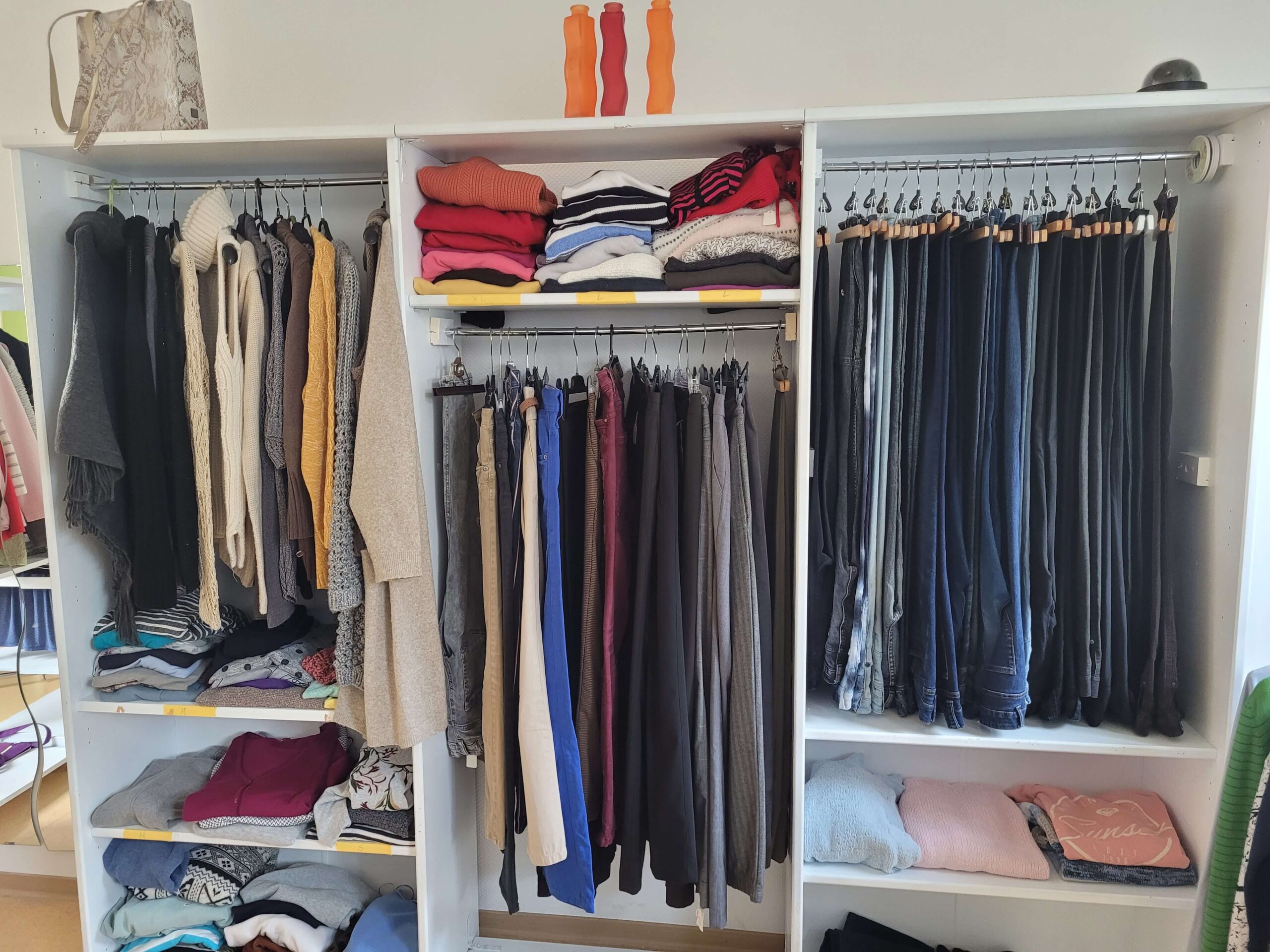 Ein weißes Regal mit aufgehängten Hose und gefalteten Pullis. Auf dem Regal stehen Vasen aus Glas