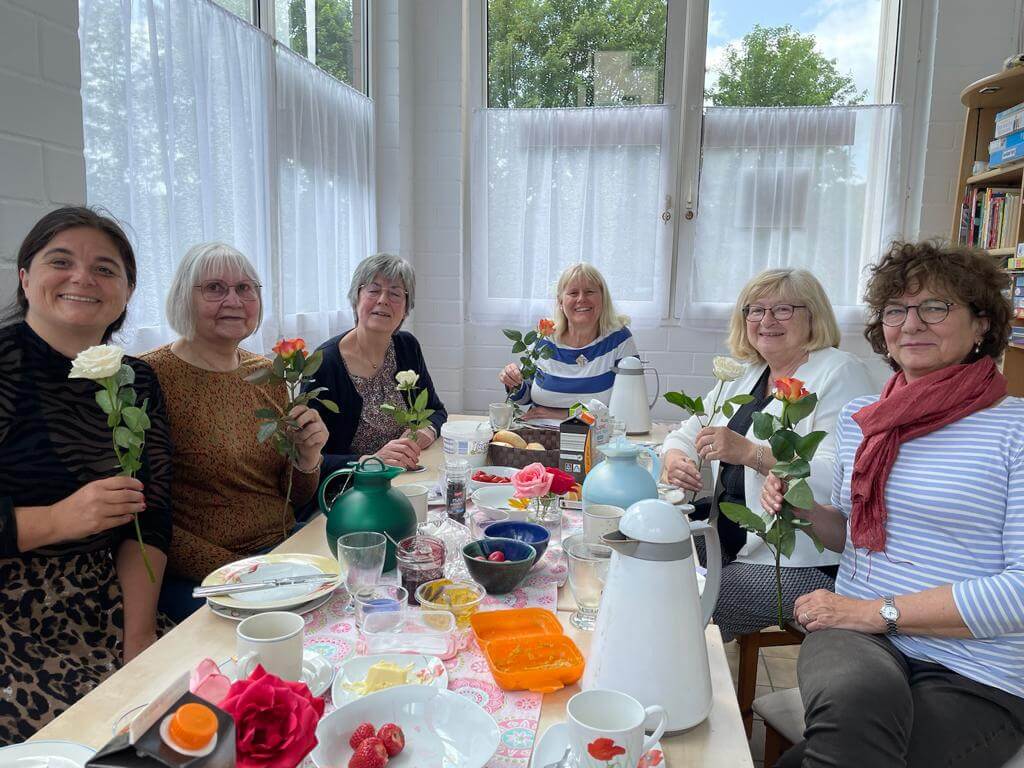 Frauen an einem gedeckten Tisch mit Rosen in der Hand, in fröhlicher Runde versammelt