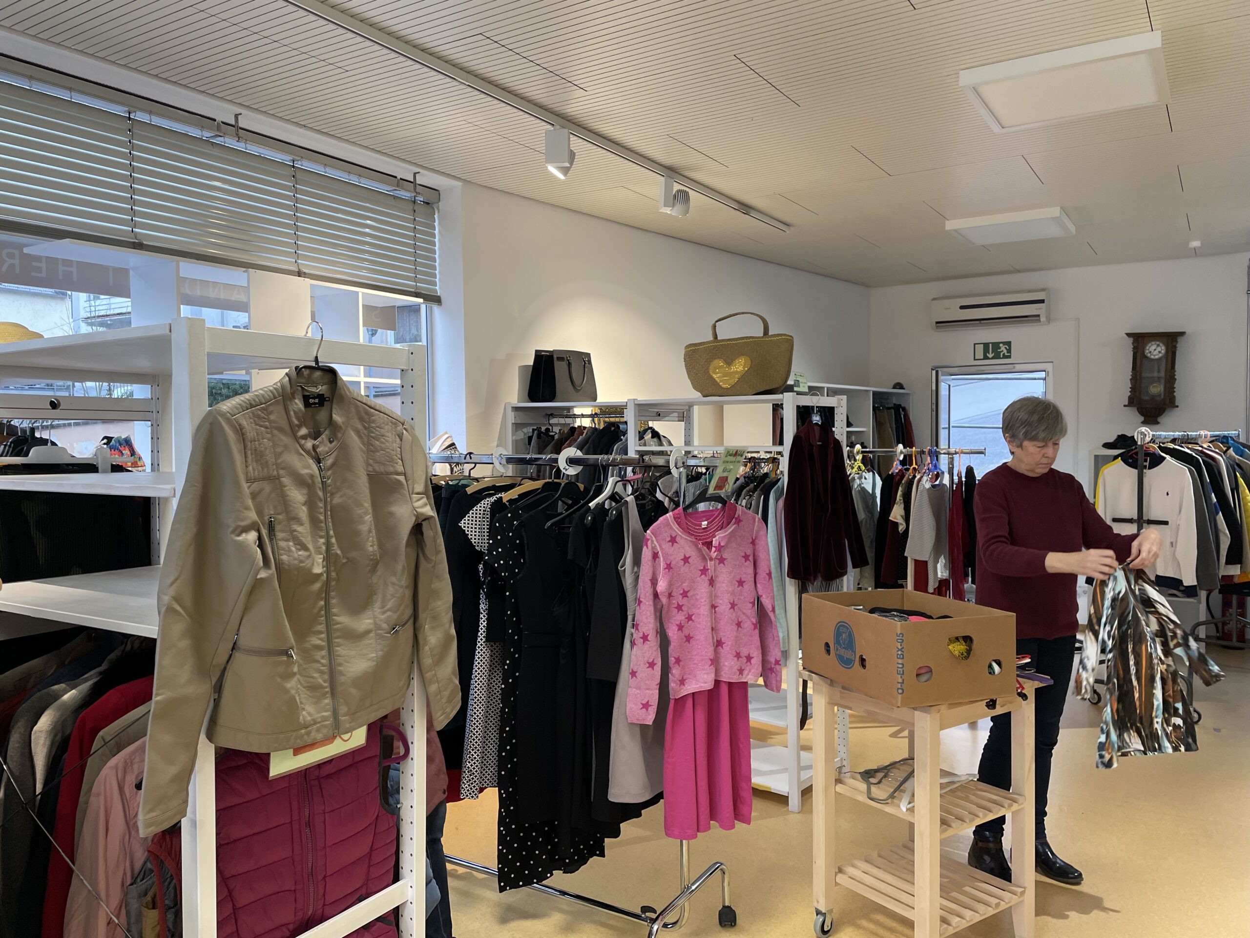 Innenraum des Second-Hand-Ladens mit zahlreichen Kleiderständern und Regalen, gefüllt mit Kleidung. Eine Mitarbeiterin faltet Kleidung zusammen.