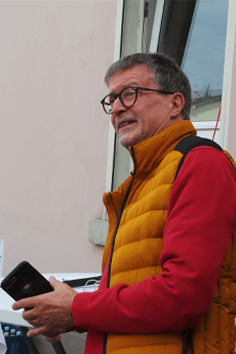 Mann mit orangefarbener Weste und Brille, hält ein Handy und schaut in die Ferne.